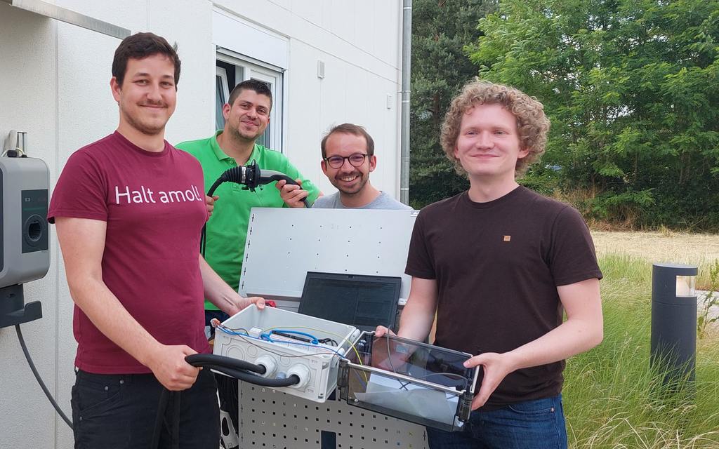 Vier Praktikanten neben einem der Forschungshäuser mit ihren Apparaturen in der Hand neben einer Wallbox. 