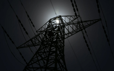 Hochspannungsmast von unten nach oben im Gegenlicht fotografiert. Die Sonne steht genau über der Mastspitze und der Strommast sowie der Himmel wirken dadurch sehr düster, die Hochspannungsleitungen sind kaum erkennbar.